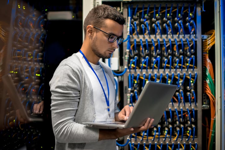 Side view portrait of young man with laptop standing by server cabinet while working with supercomputer