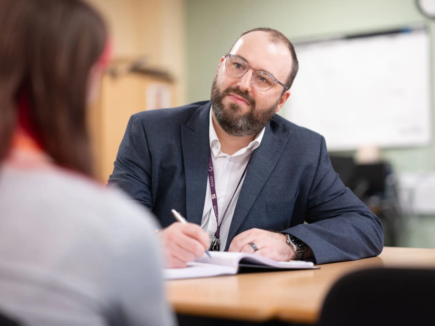 A man in a suit and glasses sits at a desk, writing in a notebook while speaking to a student.