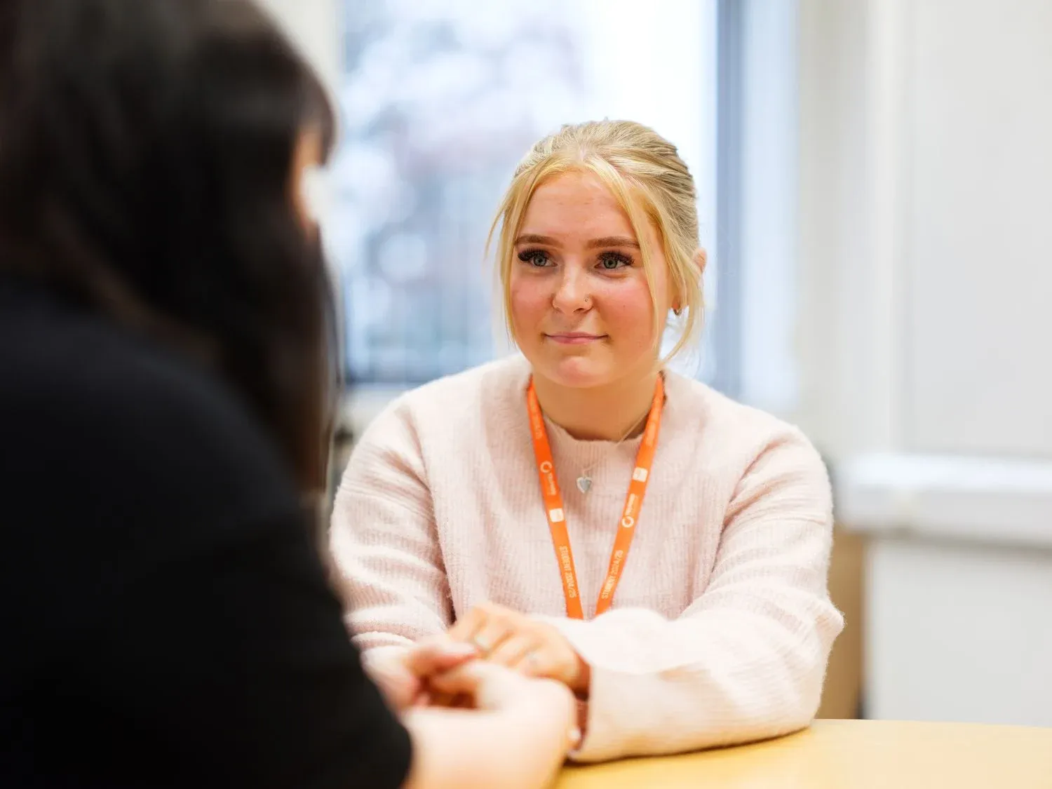 Two people sitting at a table, engaged in conversation. One wears an orange lanyard with a heart pendant.