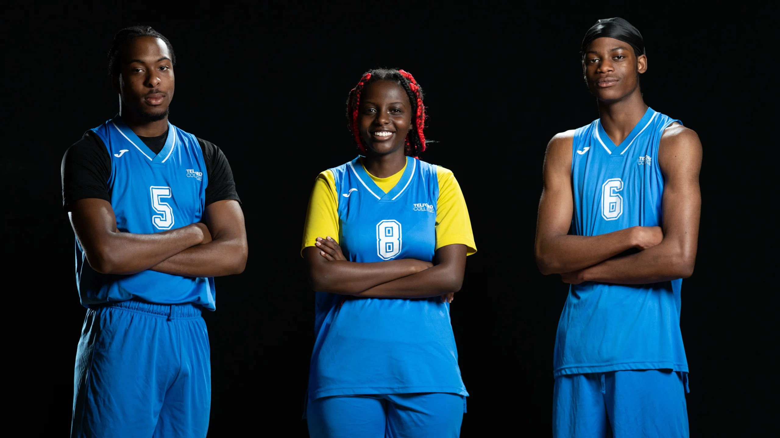 A group of six students in blue sports uniforms holding basketballs, a volleyball, and a football, posing on a wooden floor against a black background.]